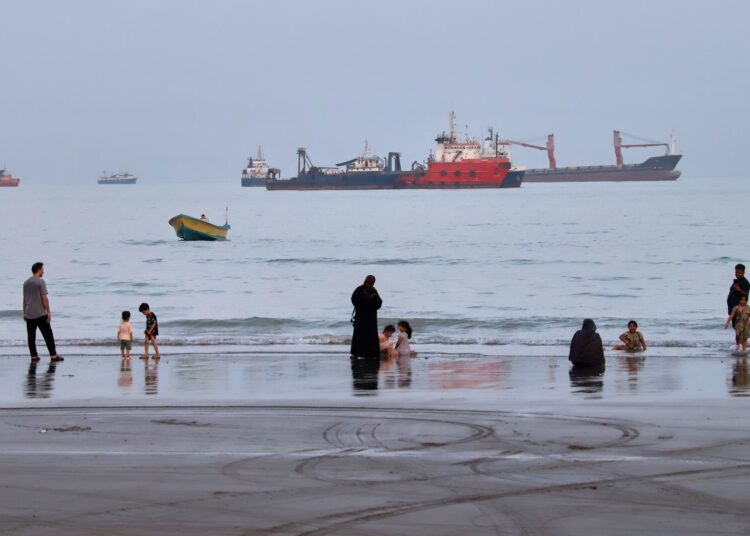 Pobrežie Hormuzského prielivu pri iránskom meste Bandar Abbas priťahuje kúpajúcich sa. V pozadí lode, čakajúce na prechod úžinou. Foto: SITA/Amirhosein Khorgooi/ISNA via AP
